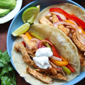 Two assembled fajitas on a blue plate with the sheet pan fajita fillings of chicken, peppers, and onions as well as sour cream as a topping. Served with lime wedges.
