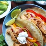 Two assembled fajitas on a blue plate with the sheet pan fajita fillings of chicken, peppers, and onions as well as sour cream as a topping. Served with lime wedges. Surrounding the plate are small dishes of salsa, sour cream, and avocado slices.